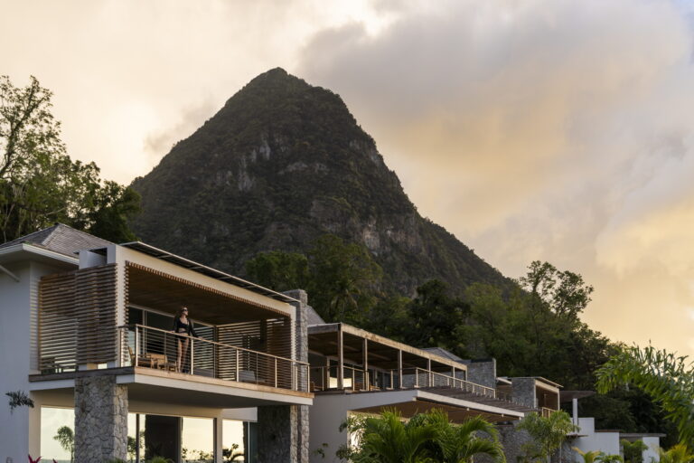 View of the Spice of Life Villa at Sugar Beach in Saint Lucia, a modern, white-and-stone mountain side property with wooden accents. A woman in black swimwear stands on the upper-level balcony, framed by lush green foliage and a towering mountain peak in the background under a soft, warm sky.
